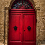 A red door with a metal handle set within a stone archway in Mdina, showcasing classic architectural details.