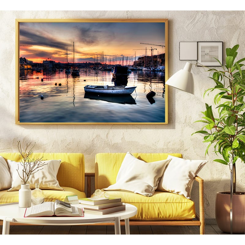 Gold frame with a photo of a boat in the Kalkara Marina during golden hours hanged above 2 yellow arm chairs with a white oval cofee table with some books on top