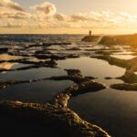 a man with a fishing rod in the distance at sunrise near saltpans in marsascala coastline with reflections