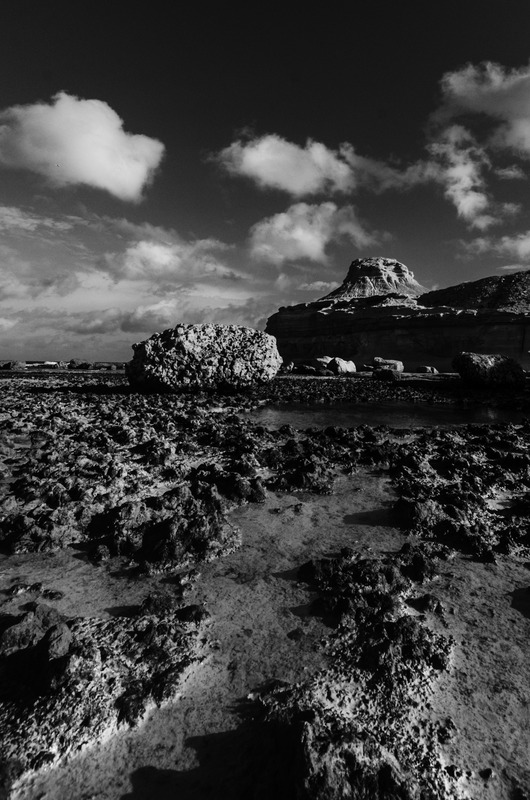 Monochrome photograph of rugged rocks and tranquil water at Gozo Qbajjar, highlighting the serene coastal landscape.