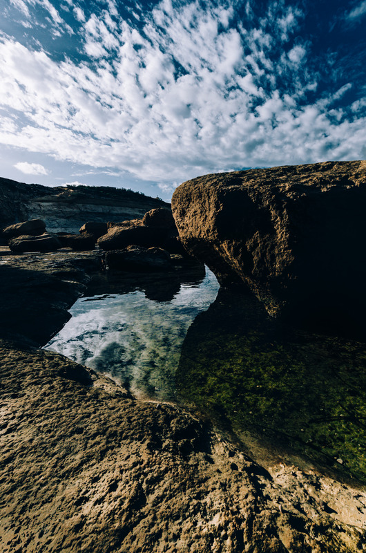 Rocky shoreline with tidal pool under a blue sky with wispy clouds at Gozo Qbajjar