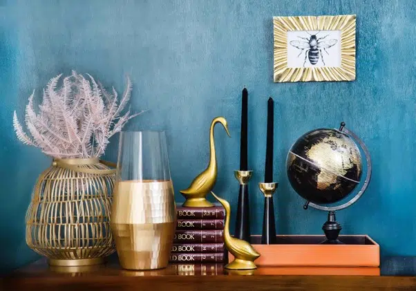 A blue wall serves as a backdrop for elegant gold vases and a decorative bird cage, creating a vibrant interior scene.