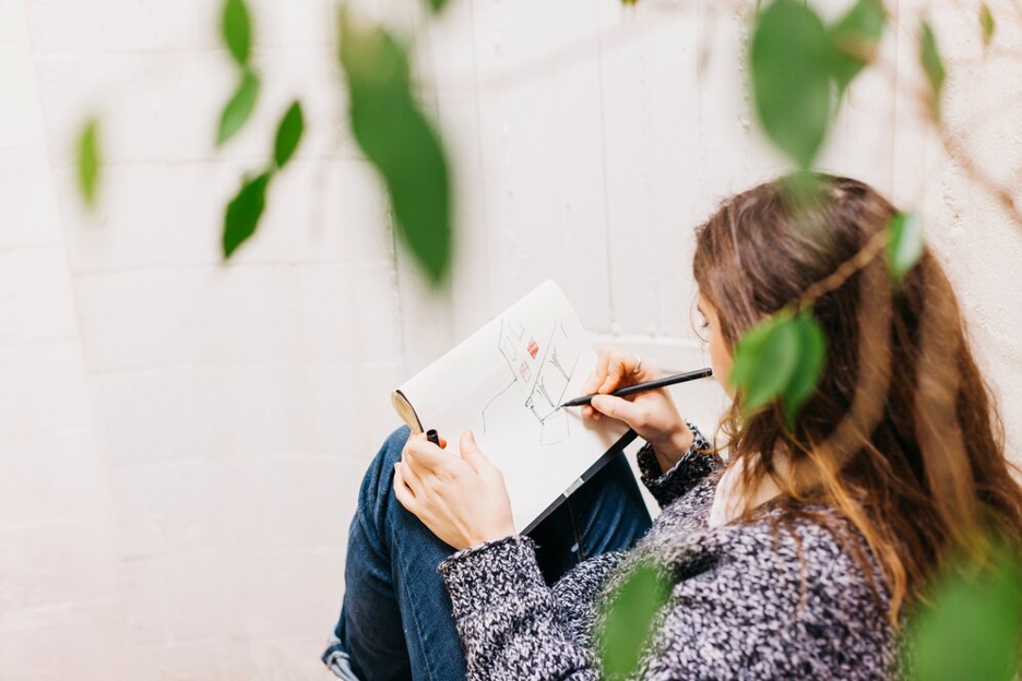 A woman sits on the ground, pen in hand, immersed in her drawing, symbolizing the breaking of creative barriers.