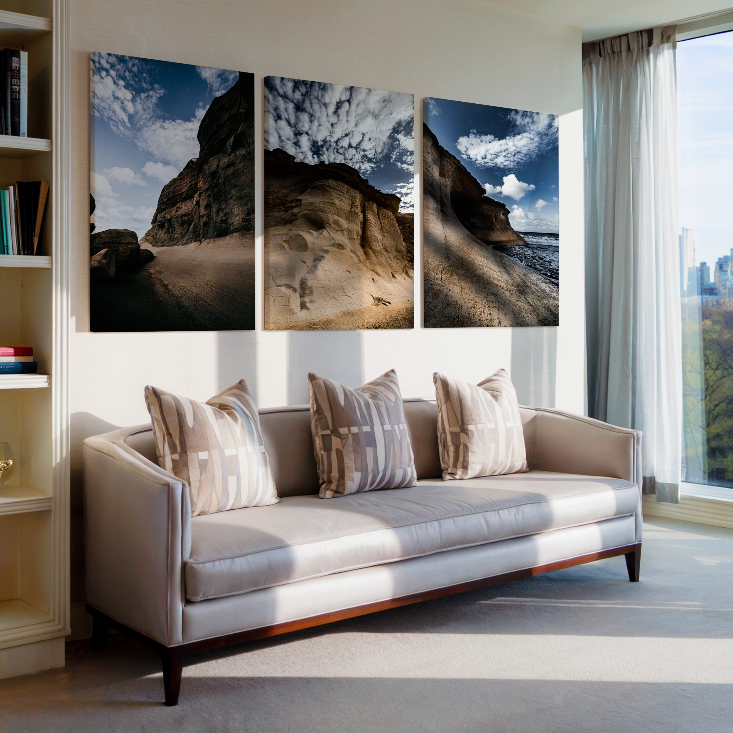 A triptych wall art set featuring dramatic rock formations and a coastal landscape in Qbajjar, Gozo. The prints are displayed above a modern beige sofa in a well-lit living room with a large window showcasing a city skyline.