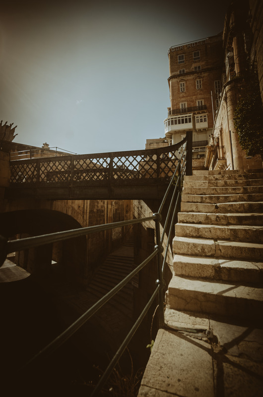 A historic iron bridge and weathered stone staircase in Valletta, Malta, bathed in warm sunlight, evoking a sense of nostalgia and timeless beauty.