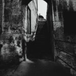 Black and white photograph of an arched stone passageway in Valletta, Malta, leading to a dimly lit staircase with a silhouetted figure at the top. The textured walls show the historic architecture of the city.