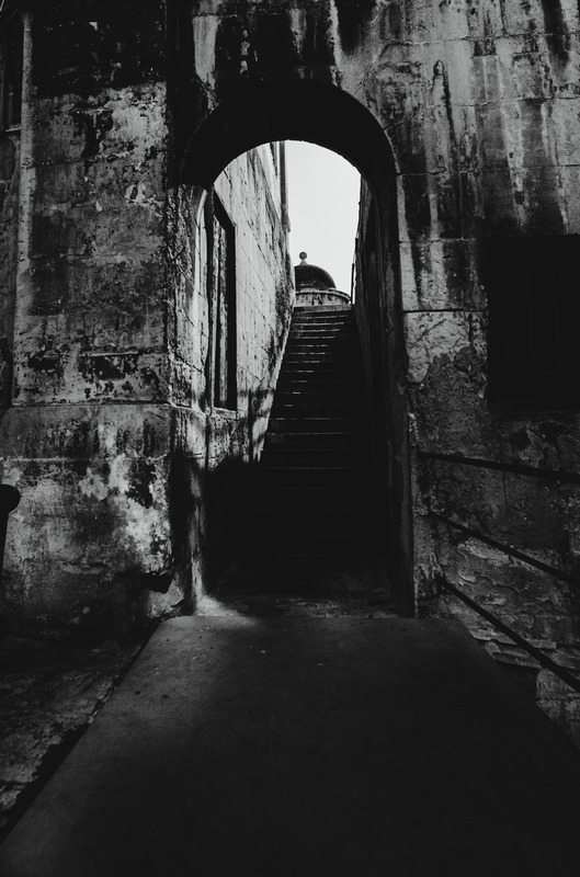 Black and white photograph of an arched stone passageway in Valletta, Malta, leading to a dimly lit staircase with a silhouetted figure at the top. The textured walls show the historic architecture of the city.