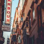 A vintage Maltese street scene in Valletta featuring old signage, traditional balconies, and warm architectural tones under a clear blue sky.