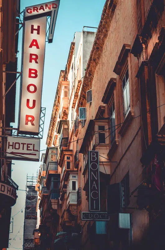 A vintage Maltese street scene in Valletta featuring old signage, traditional balconies, and warm architectural tones under a clear blue sky.