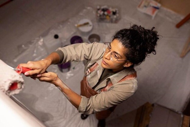 A woman applies paint to a white wall with a brush, representing the theme of using real estate to finance her college education.