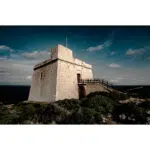 Historic watchtower under a dramatic sky, surrounded by green shrubs and overlooking the sea.