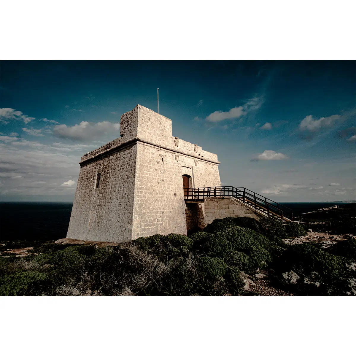 Historic watchtower under a dramatic sky, surrounded by green shrubs and overlooking the sea.