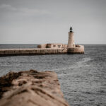 Lighthouse on the Valletta breakwater, seen across the sea with a textured stone wall in the foreground and a muted sky.