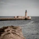 Lighthouse on the Valletta breakwater, seen across the sea with a textured stone wall in the foreground and a muted sky.