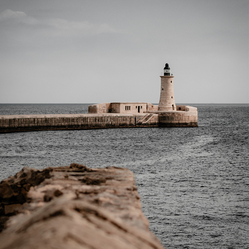 Lighthouse on the Valletta breakwater, seen across the sea with a textured stone wall in the foreground and a muted sky.