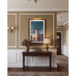 A minimalist living room with a large window, a green sofa, and a natural wood coffee table. On the wall above the sofa, a framed print of the Gozo Xewkija Il-Mitħna tat-Tmien Kantunieri artwork is displayed.