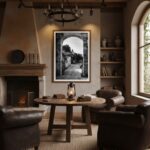 A large, framed print of the Malta Mtahleb Church Archway hanging on a light grey living room wall above a brown leather sofa with a white cushion and a soft grey throw blanket. To the left of the sofa is a brown wooden side table with a green plant in a white pot and a stack of books. A window with natural light is visible in the background.