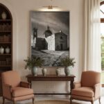 A framed print of Malta Mtahleb Church hangs on a light blue wall above a white sofa with neutral throw pillows, a woven blanket, and a small wooden side table with a plant, creating a serene living room atmosphere.