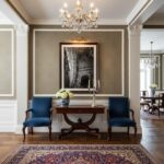 A large, framed print of the Mgarr Malta Church Arch hangs on a light grey wall in a modern living room, above a minimalist off-white sofa. A decorative throw blanket and throw pillow add texture to the sofa. To the left of the sofa, a tall, leafy green plant in a terracotta pot adds a touch of nature. The room is well-lit, suggesting natural light, and the overall impression is clean, serene, and stylish. The artwork itself features the distinctive architecture of the Mgarr Malta Church, with its prominent arch, under a bright sky.