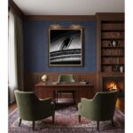 A large, brightly lit living room with a cozy, minimalist aesthetic. The room features a light gray sofa with a wooden frame and white cushions, a simple wooden coffee table with a plant, and a large window looking out onto greenery. On the wall above the sofa hangs a large, framed print depicting the Mgarr Malta Church Blocks in a modern art style.