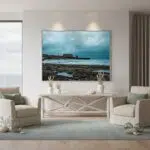 A person wearing a blue t-shirt stands on a rocky beach at dusk, looking out at the sea where a historic battery is silhouetted against the colorful sky.