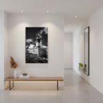 A person stands on a balcony overlooking the sea, holding a framed print of the Malta Marsaxlokk Lighthouse in black and white, with a minimalist interior design and natural light.