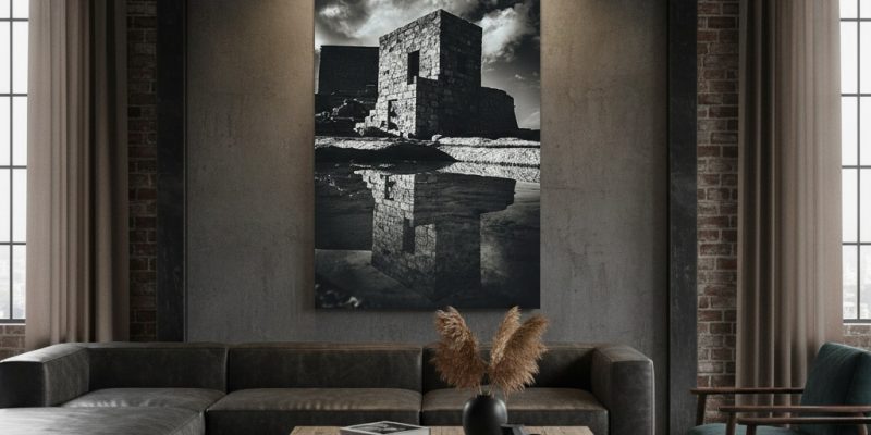 A modern, minimalist living room with a large window looking out onto a cityscape. A person is sitting on a sofa, reading a book. The "Gozo Qbajjar Battery Monochrome - Wall Art" is hanging on the wall above the sofa, adding a touch of sophistication to the space.