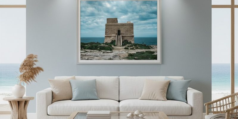 A person stands on a rocky beach at dusk, gazing at the Dwejra Tower under a twilight sky. The scene is illuminated by the soft glow of the blue hour, highlighting the tower's historic architecture and the rugged coastline.